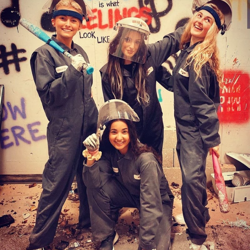 a group of friends posing for a photo inside of the rage room at chaos factory