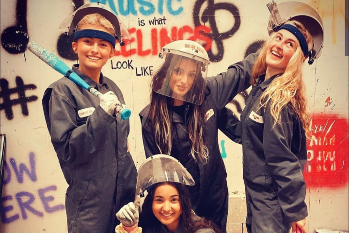 Four people in coveralls and helmets pose in front of graffiti wall with baseball bats.
