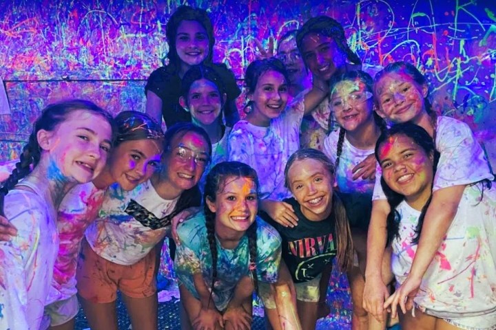 Group of kids in colorful, paint-splattered clothes smiling under blacklight.