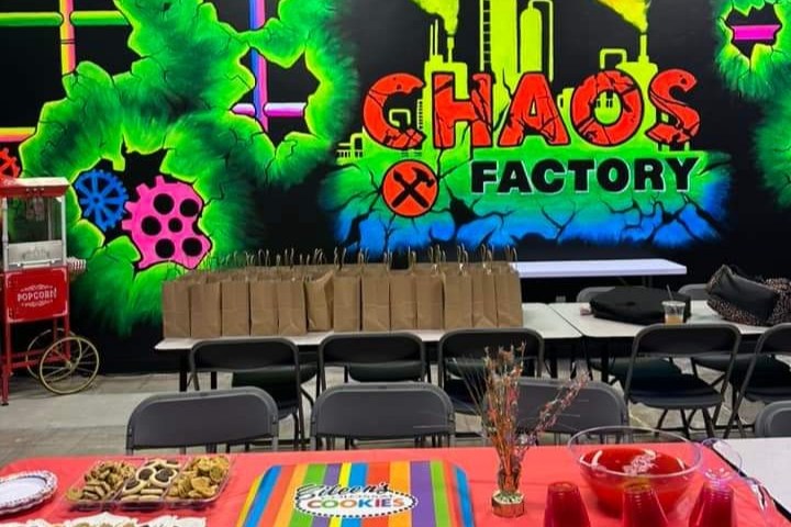 Neon wall with 'Chaos Factory' and tables with snacks and goody bags in front.
